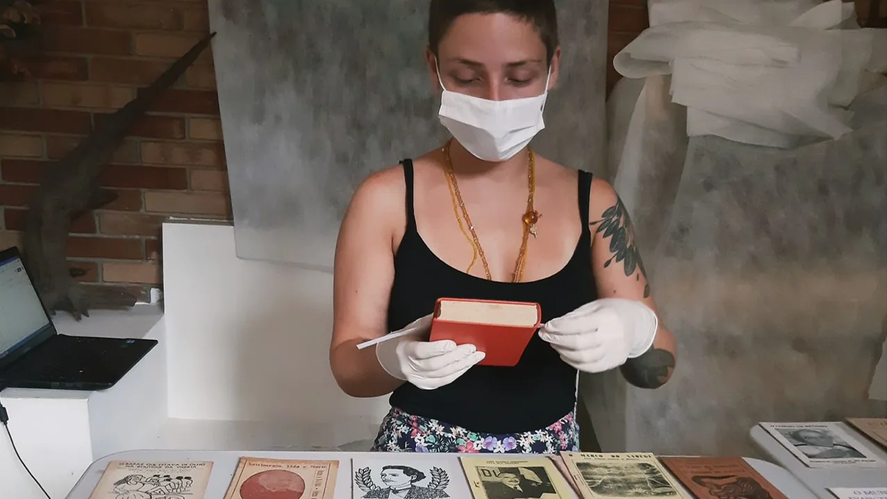 Marina Nabuco examining a small red cordel chapbook while wearing gloves and a protective mask